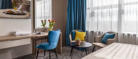 A hotel room with a bed, a desk with a tray of breakfast, a chair, a small table, and a window with curtains.
