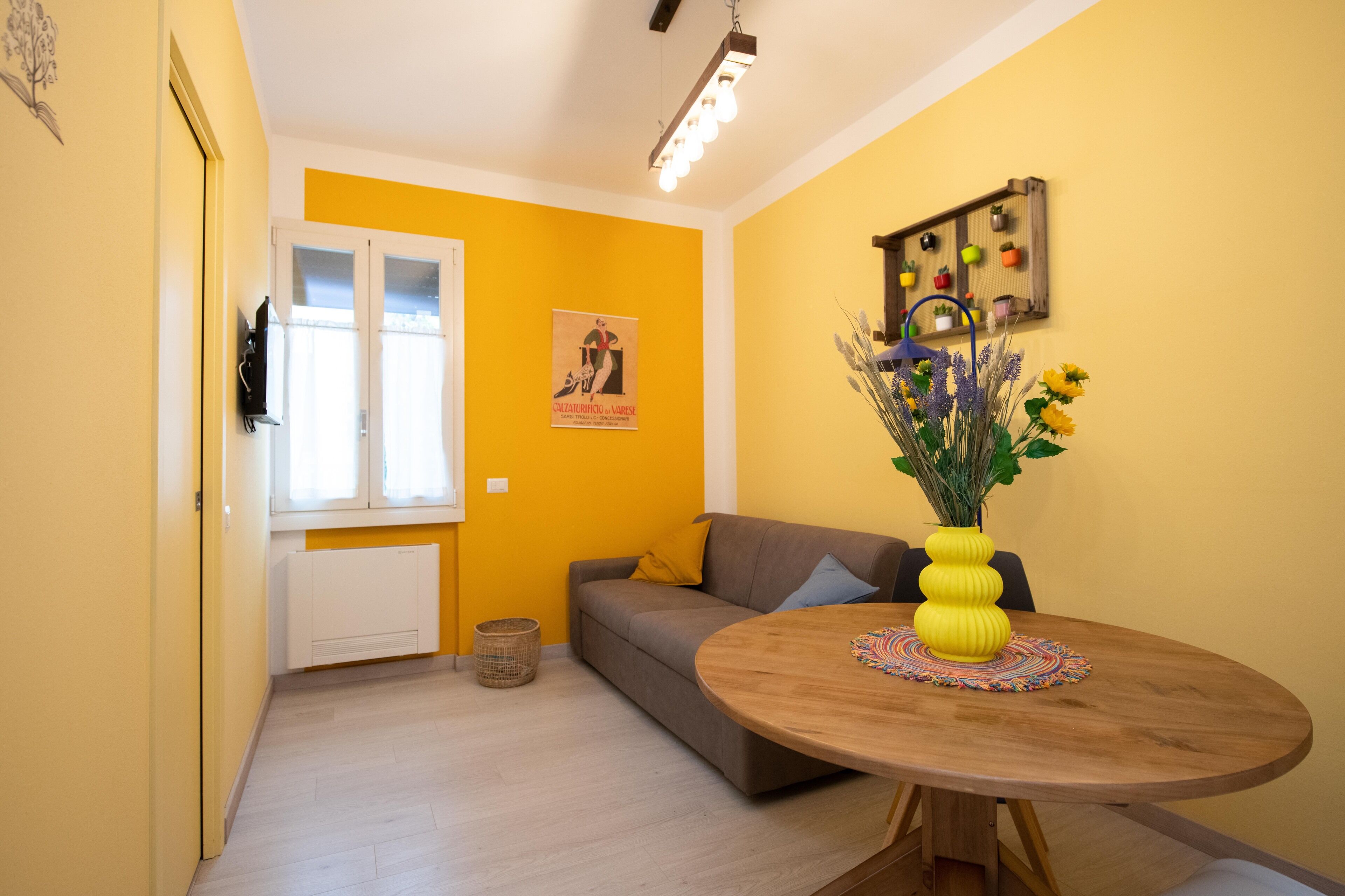 Quarto com parede amarela, sofá cinza, mesa de madeira com um vaso amarelo e um cartaz na parede.