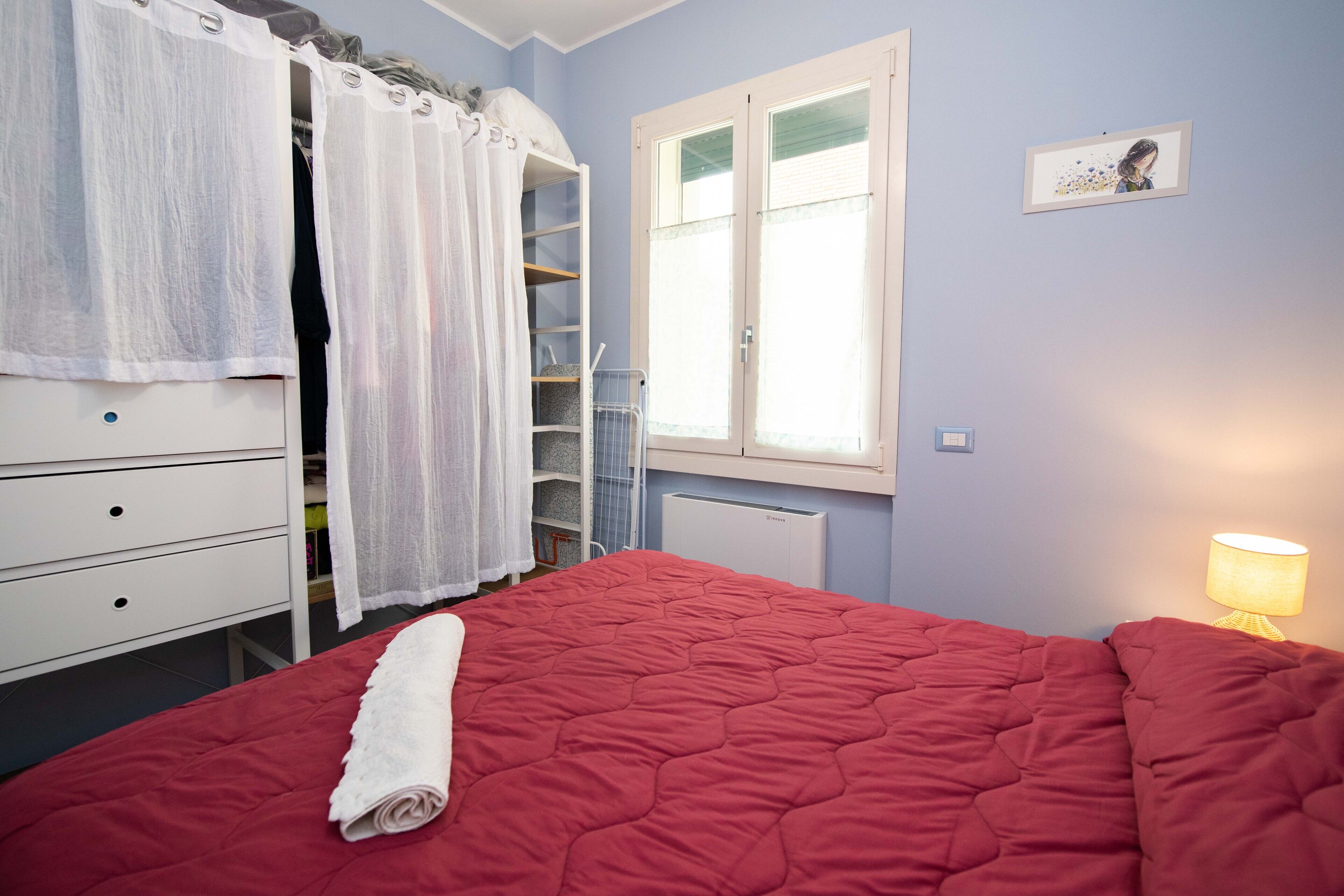 A bedroom with a red bed, white curtains, a radiator, a white dresser, and a window with a picture on the wall.