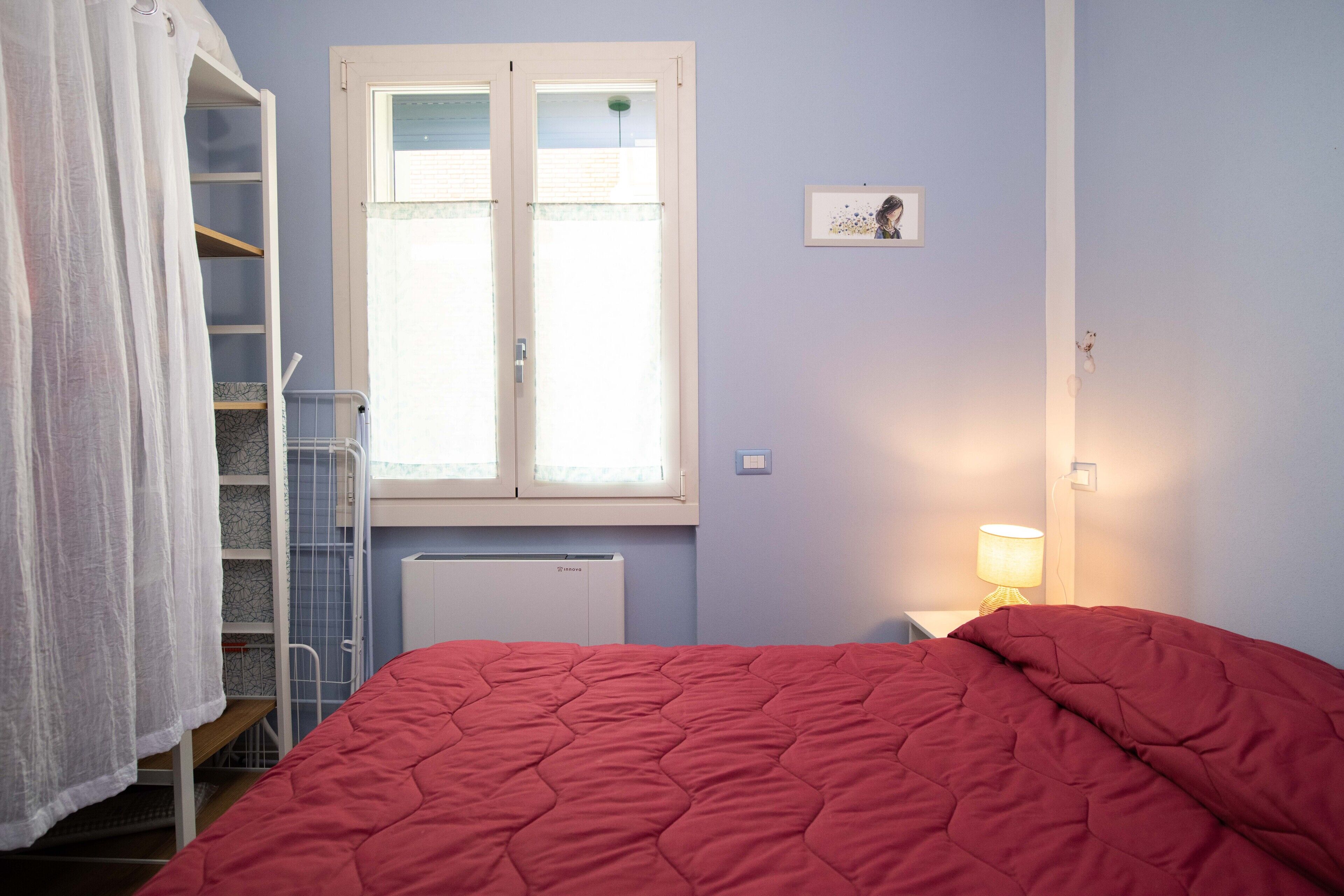 Een slaapkamer met een rode sprei, een witte radiator, een raam met zonwering, een bedlampje en een klein lijstwerk met een afbeelding aan de muur.