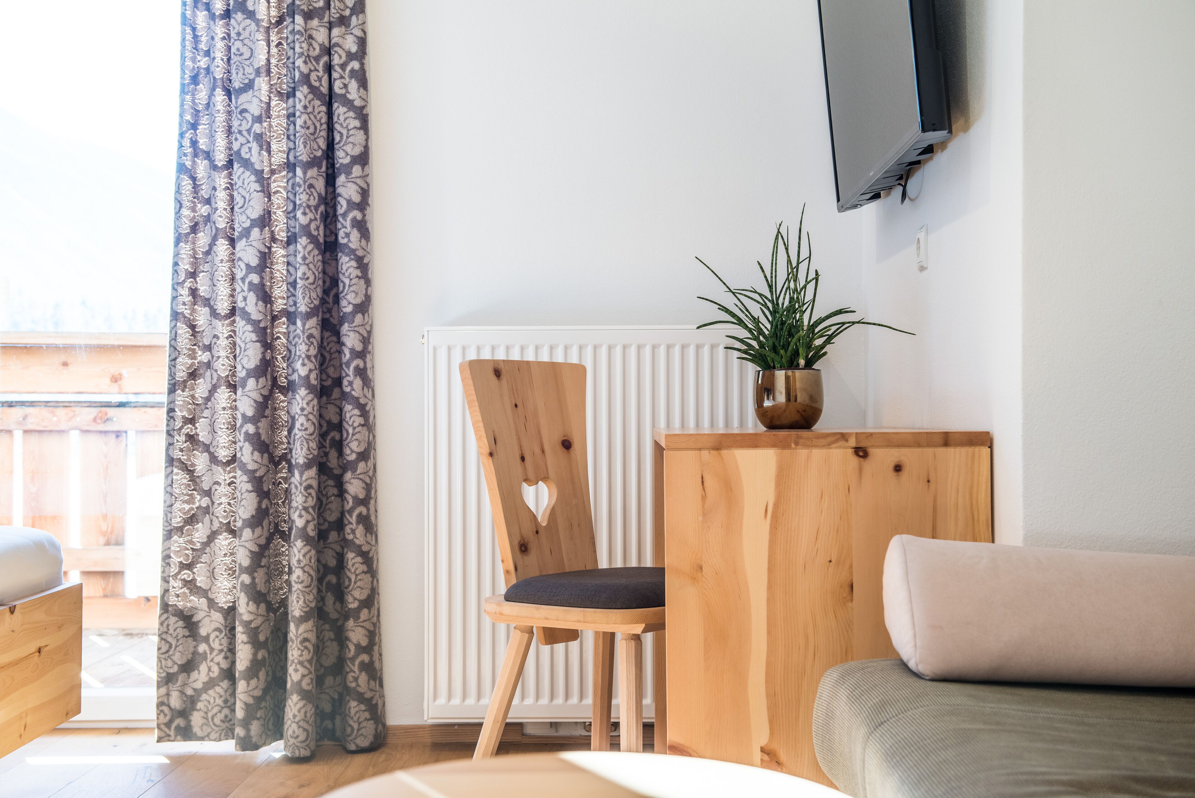 A cozy room with a wooden chair, a small table with a plant, a sofa, and a TV mounted on the wall.