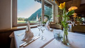 A dining table set with white napkins, silverware, and wine glasses, with a vase of yellow flowers and a view of a mountain and church through the window. - Hotel Antholzerhof (Rasun Anterselva)