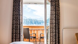 A bedroom with a bed, a window with curtains, a view of a snowy mountain landscape, and a balcony with chairs. - Hotel Antholzerhof (Rasun Anterselva)