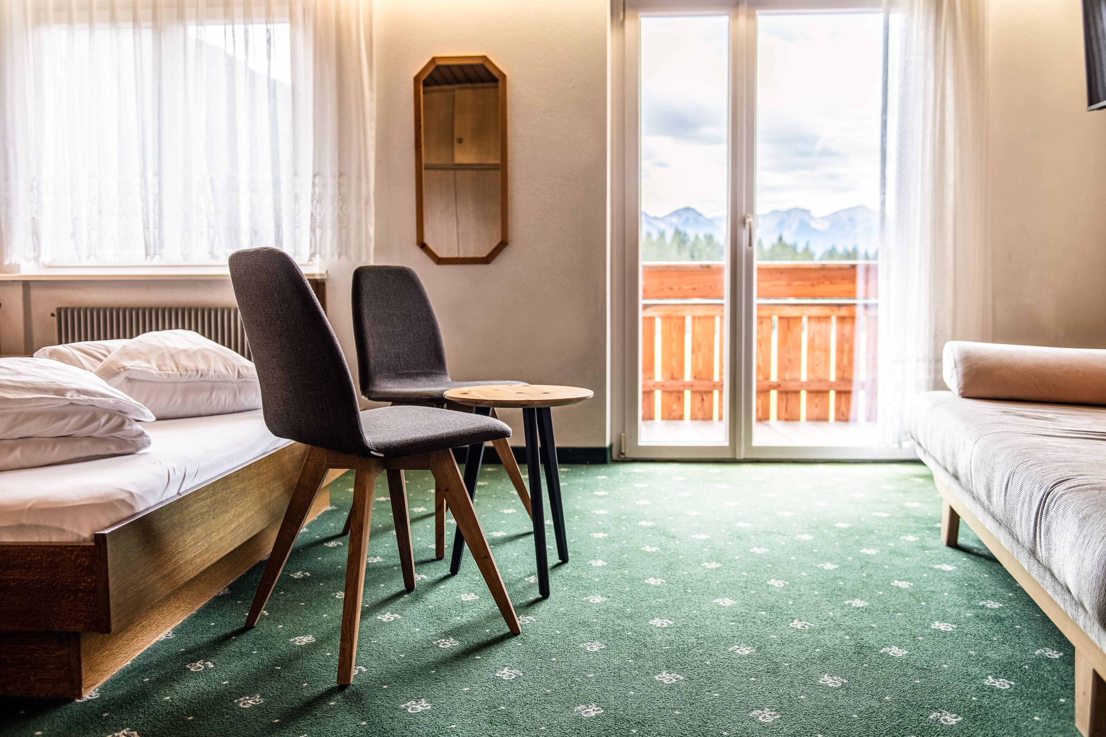A room with a bed, a chair, a small table, and a sofa. There is a window with a view of mountains and a balcony with a wooden fence.