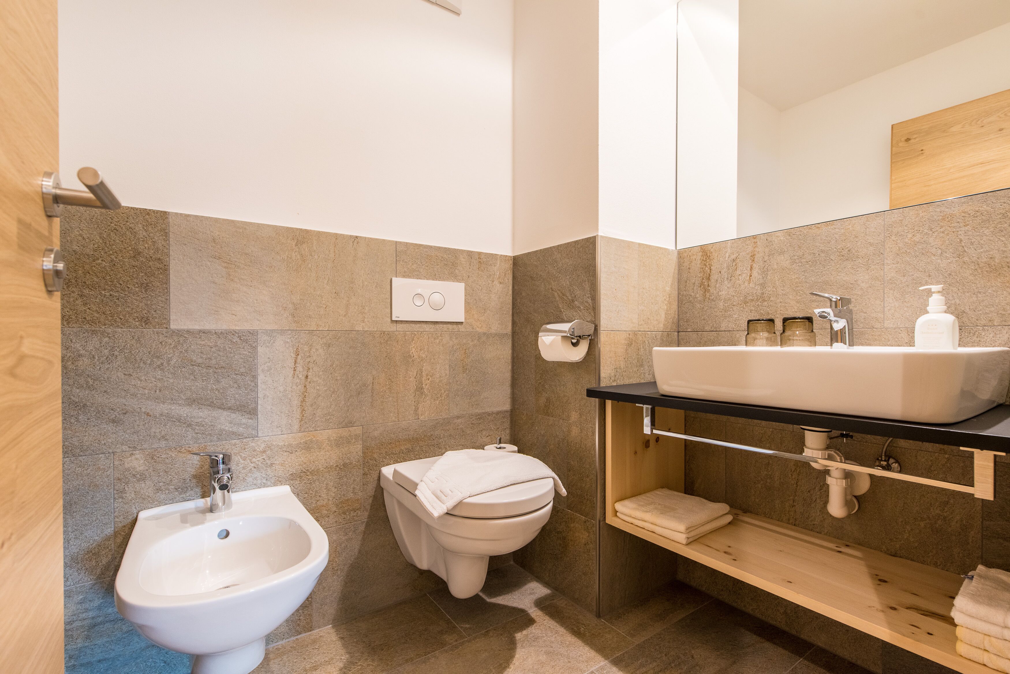 A modern bathroom with a bidet, toilet, sink, and wooden shelves.