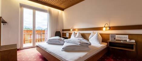 Habitación de hotel con techo de madera, una cama grande con ropa blanca, una mesita de noche con lámpara, vistas a las montañas a través de la ventana y piso cubierto de alfombra roja.