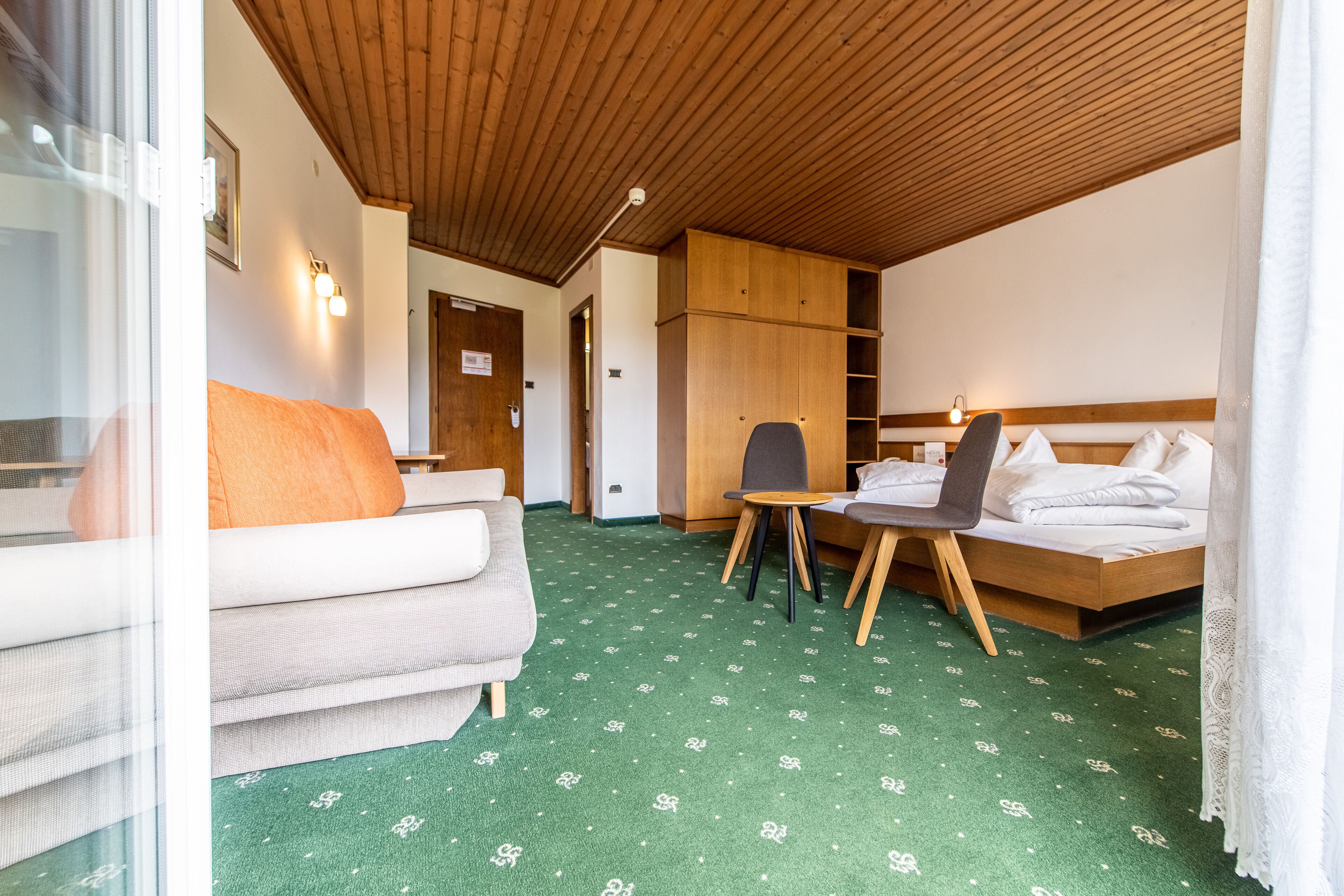 Ein Hotelzimmer mit grünem Teppich, Holzboden, einem Bett, einer Couch, einem Sessel und einem kleinen Tisch.