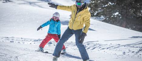 A snowy slope with a person skiing and a child in a blue jacket.