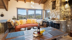 Une pièce chaleureuse avec une cheminée en pierre, une table en bois avec des sets de table, un canapé avec des coussins jaunes et une vue sur les montagnes à travers de grandes fenêtres.