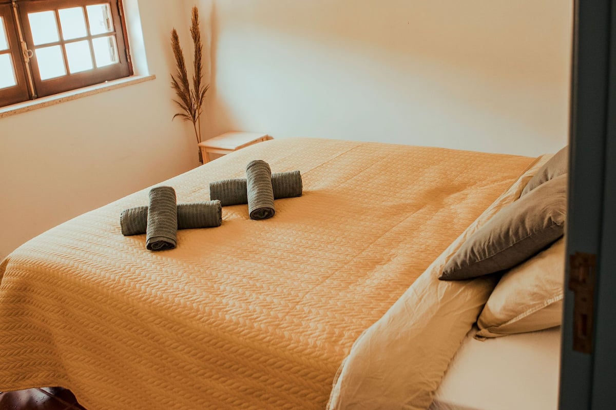 A neatly made bed with rolled towels, a window with a view, and a wall-mounted mirror.