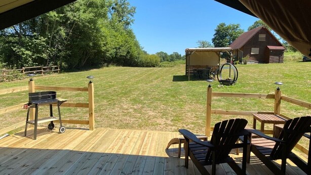 An outdoor deck with wooden chairs and a grill cart, overlooking a grassy field and a barn.
