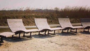 Eine Reihe aus Holzgestellten Liegestühlen mit Metallrahmen, auf einem Sandstrand mit hohem Gras im Hintergrund.
