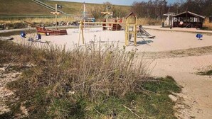 Ein Sandstrand mit Spielgeräten, einer kleinen Holzkonstruktion und einem Rettungsschwimmturm im Hintergrund.