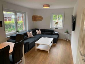 A modern living room with a black sofa, white coffee table, and a TV mounted on the wall. There are two windows providing natural light and a view of greenery outside.