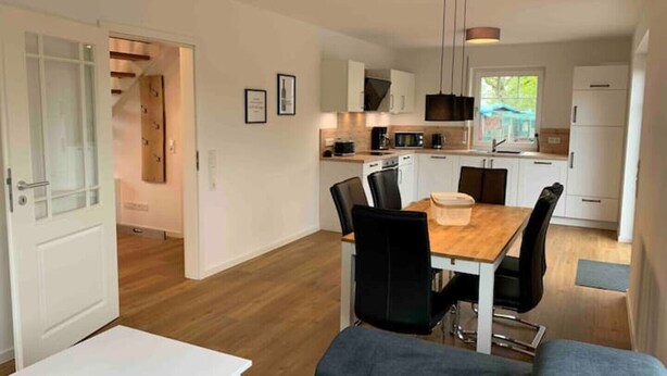Eine moderne Küche mit weißen Schränken, einem Essbereich mit Holztisch und schwarzen Stühlen sowie einer sichtbaren Tür zu einem weiteren Raum.