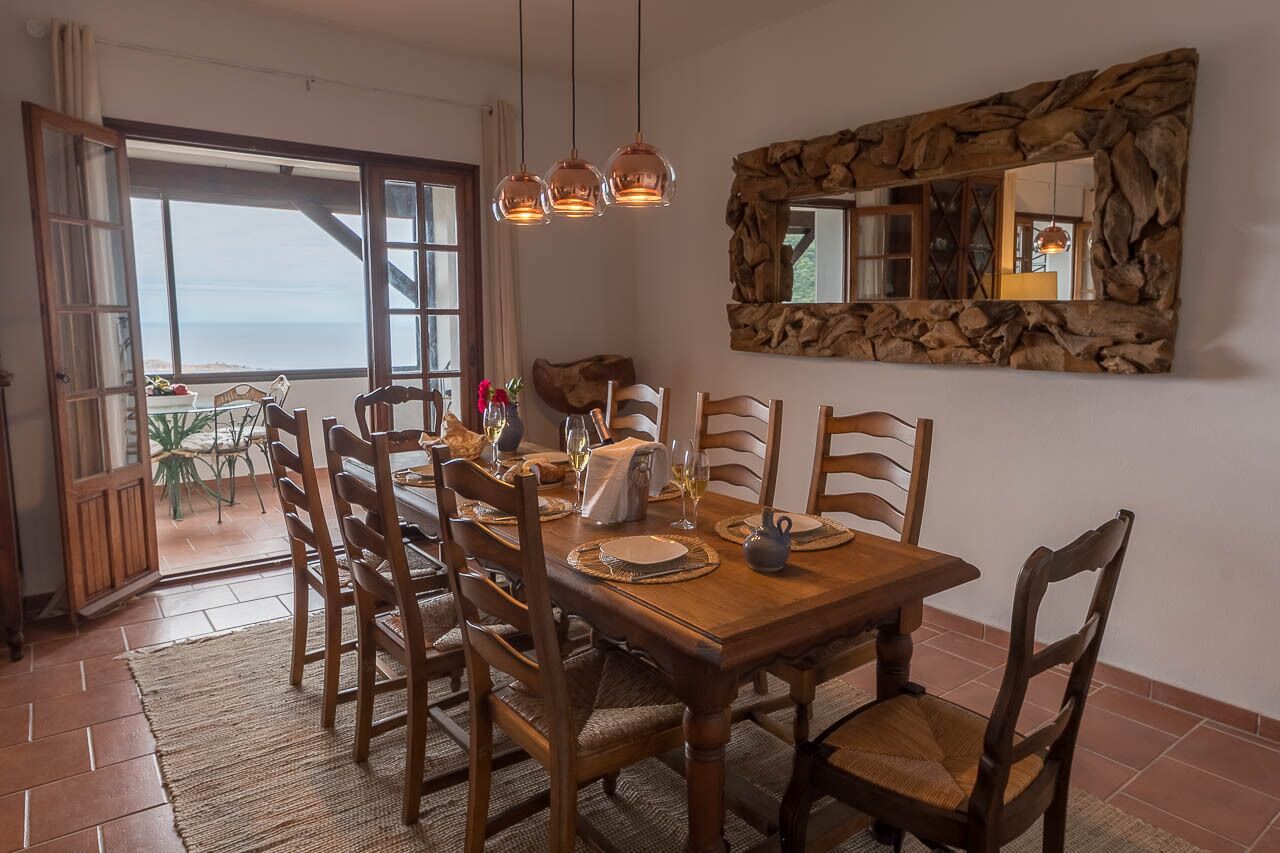 Een eetgedeelte met een houten tafel en stoelen, een spiegel met stenen lijst, en uitzicht op zee door een groot raam.