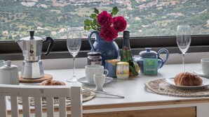 Una mesa servida con una cafetera, tetera, copas de vino y un croissant, con vistas a un paisaje costero.