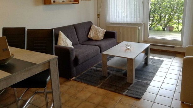 A living room with a sofa, a coffee table, a dining table with chairs, a radiator, a window with a view of greenery, and wall-mounted shelves with decorative items.