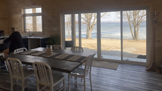 A cabin with a dining area, a kitchen, and large windows offering a view of a lake and trees.