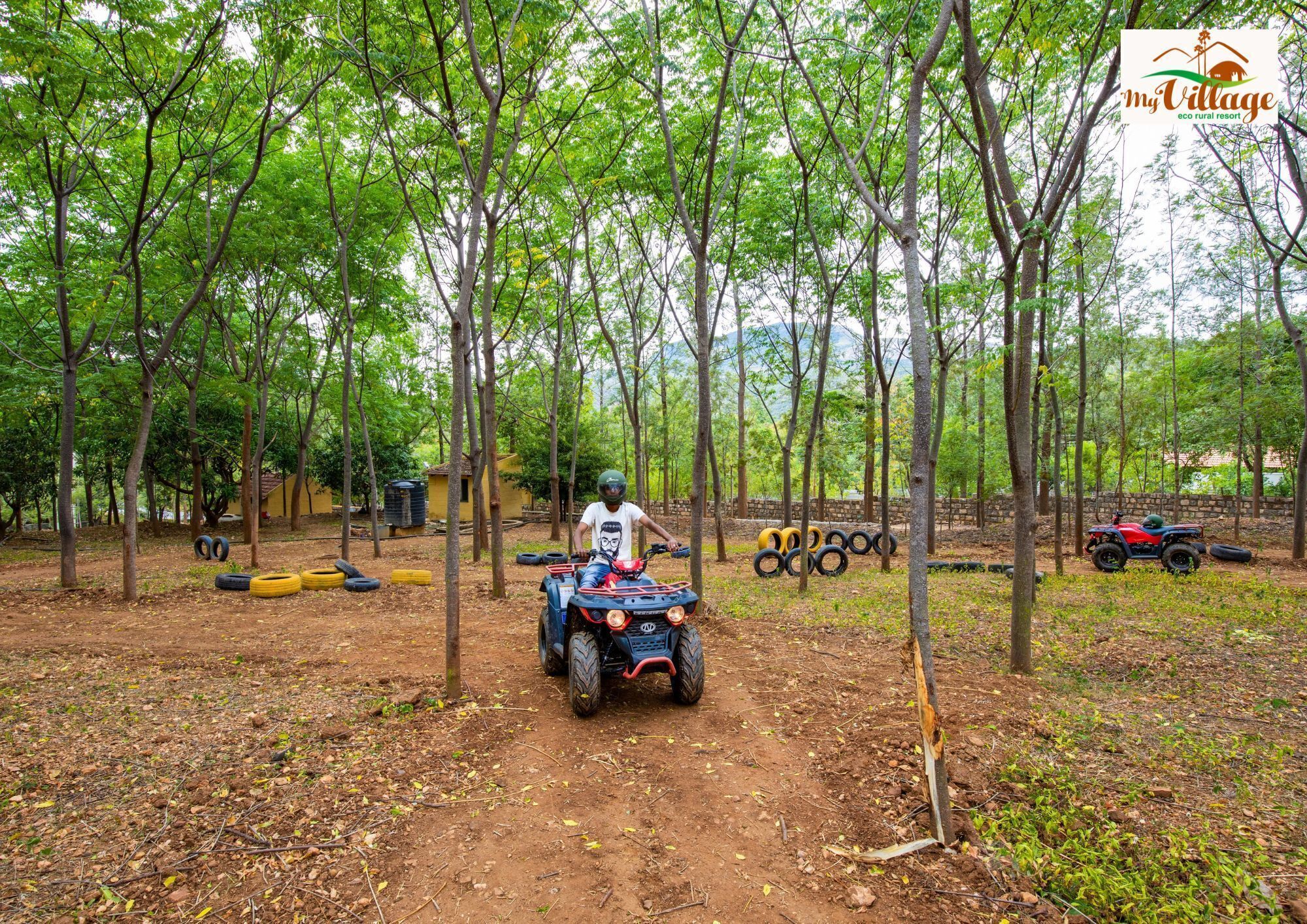 A dirt path in a forested area with a person on a quad bike, surrounded by trees and scattered tires.