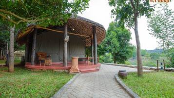 A rustic outdoor seating area with a thatched roof, surrounded by lush greenery and a paved pathway.