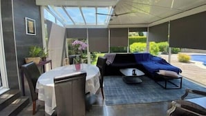 Een zonnedoek met een glazen dak, een eettafel met stoelen, een bank en een kleine tafel met een plant in pot.