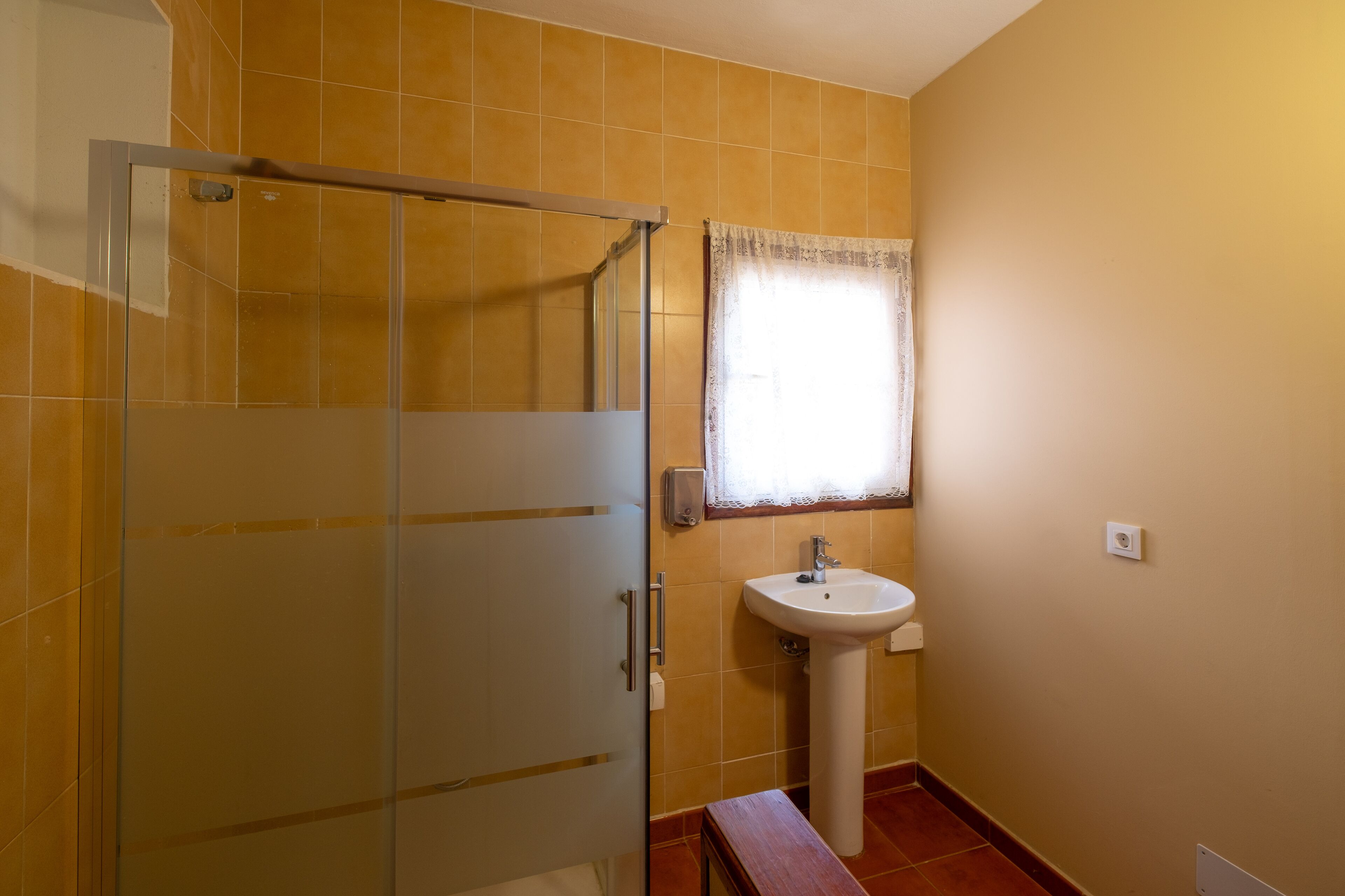 A bathroom with a glass shower enclosure, a white sink, and a window with a lace curtain.