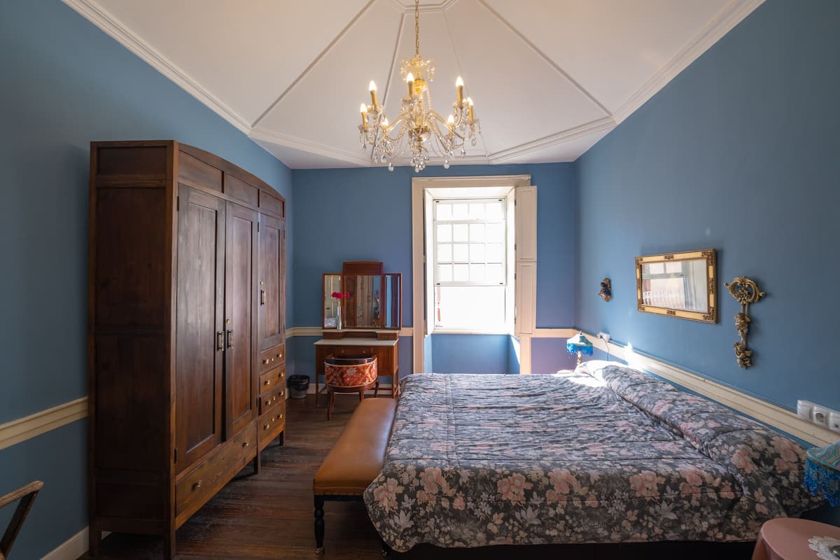 A bedroom with a large wooden wardrobe, a floral bedspread, a bench, a desk, and a chandelier.