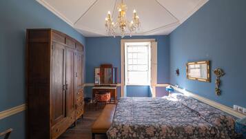 A bedroom with a large wooden wardrobe, a floral bedspread, a bench, a desk, and a chandelier.