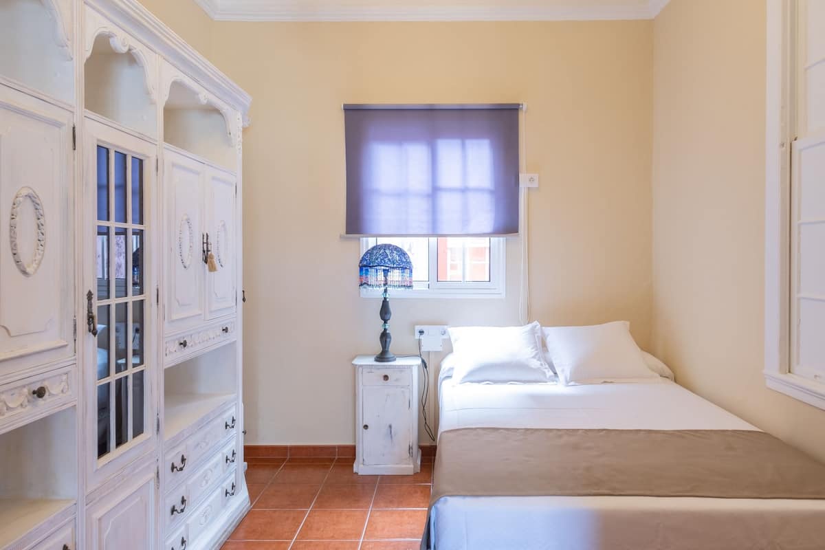 A bedroom with a bed, a nightstand, a lamp, a wardrobe, and a window with a blind.