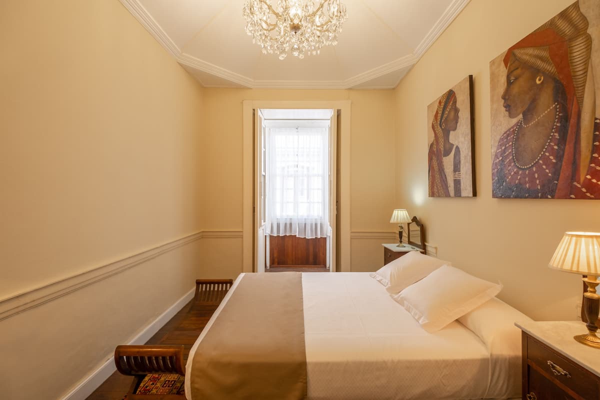 A hotel room with a large bed, a chandelier, a nightstand with a lamp, and two paintings on the wall.