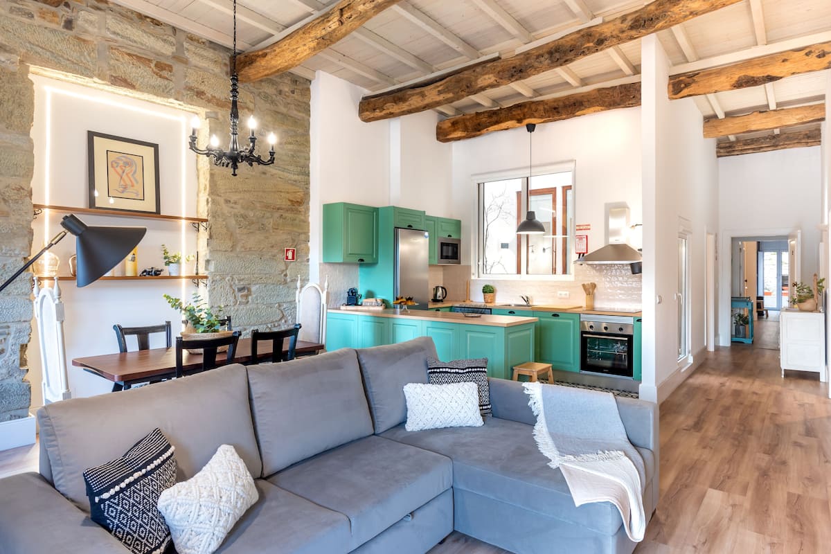 A modern living room with a grey sofa, a dining area with a wooden table, and a kitchen with teal cabinets and a central island.