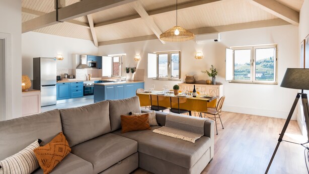 Ein modernes Wohnzimmer mit einem grauen Ecksofa, einem Essbereich mit Holztisch und einer Küche im Hintergrund.