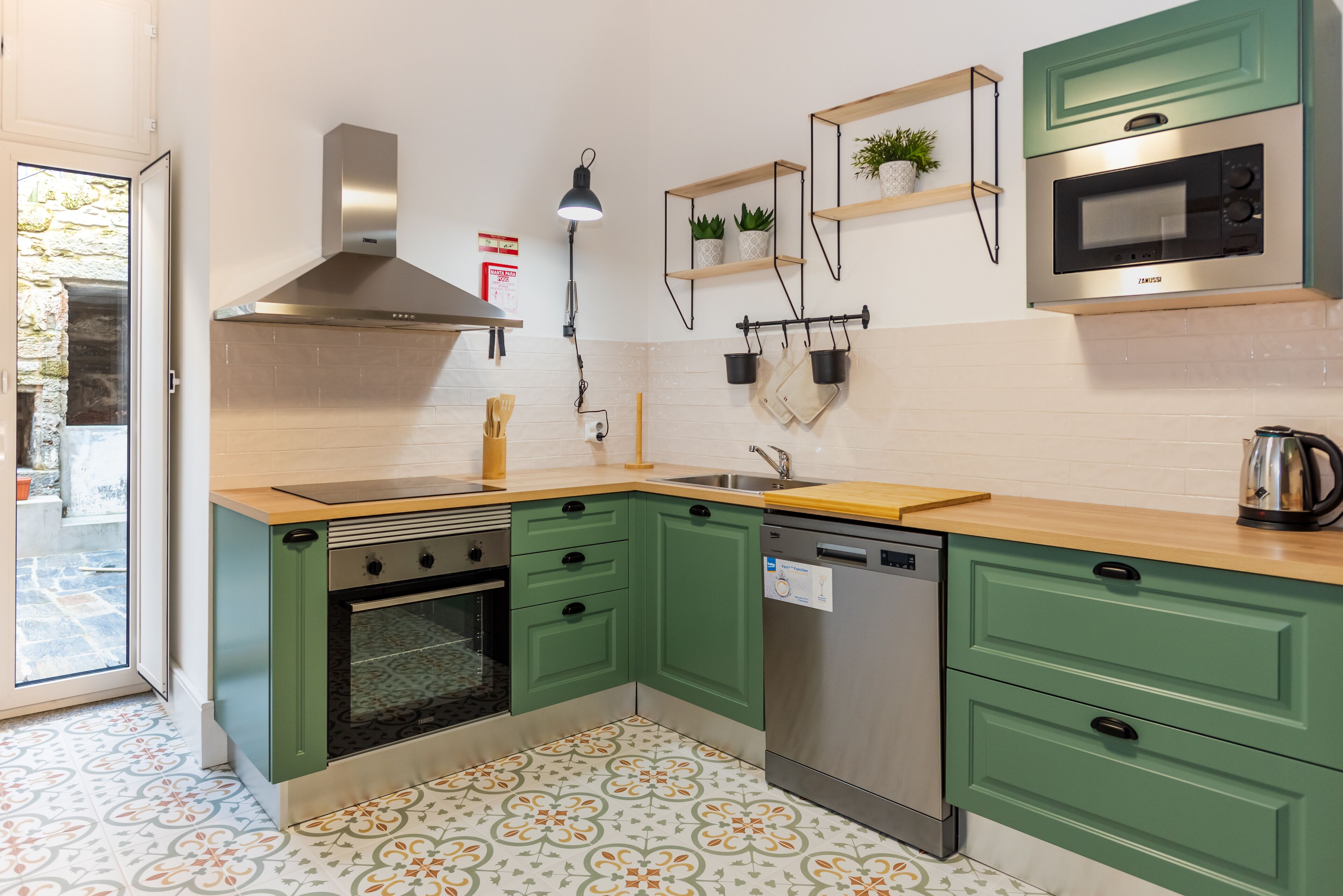 Une cuisine moderne avec des armoires vertes, une hotte de cuisinière en acier inoxydable, un four encastré, un lave-vaisselle et un four à micro-ondes. On y trouve des étagères en bois ornées de plantes, un luminaire suspendu et un sol à motifs.