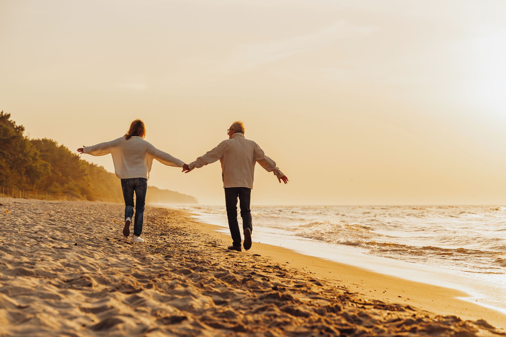 Zwei Personen gehen Hand in Hand am sandigen Strand, im Hintergrund das Meer.