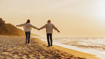 Zwei Personen gehen Hand in Hand am sandigen Strand, im Hintergrund das Meer.