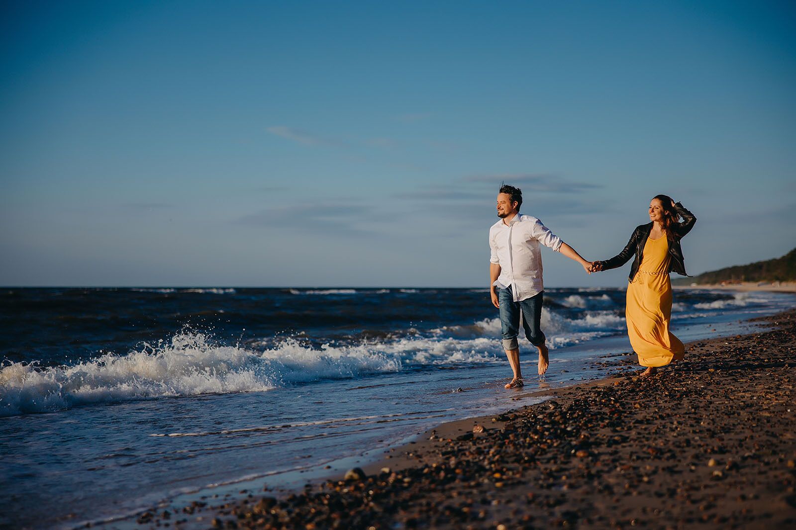A couple walking on a pebbly beach with the ocean in the background.