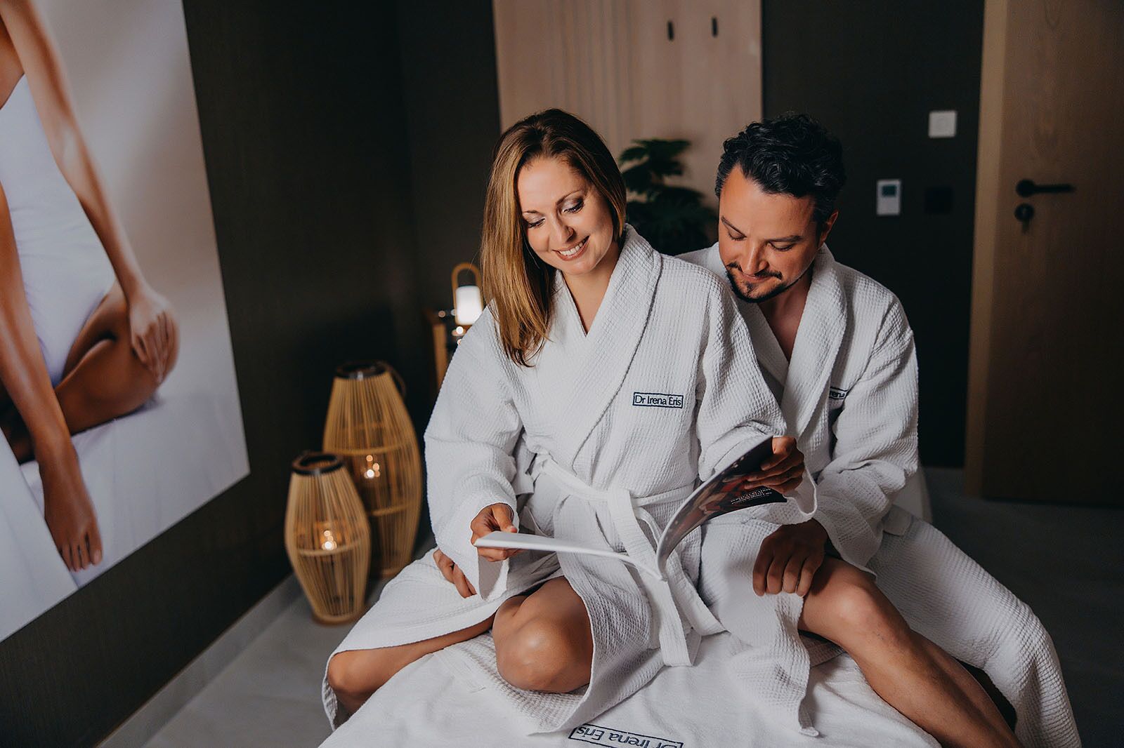 Ein Paar in einem Spa-Ambiente, beide in weiße Bademäntel gehüllt, mit einem Poster für eine Spa-Behandlung an der Wand dahinter.