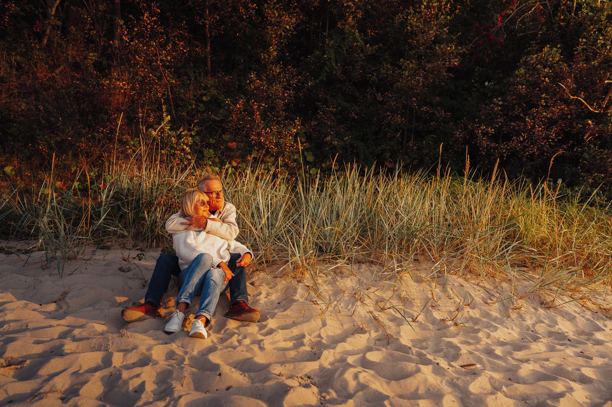 Un couple est assis sur une plage de sable, avec de hautes herbes et des buissons en arrière-plan.