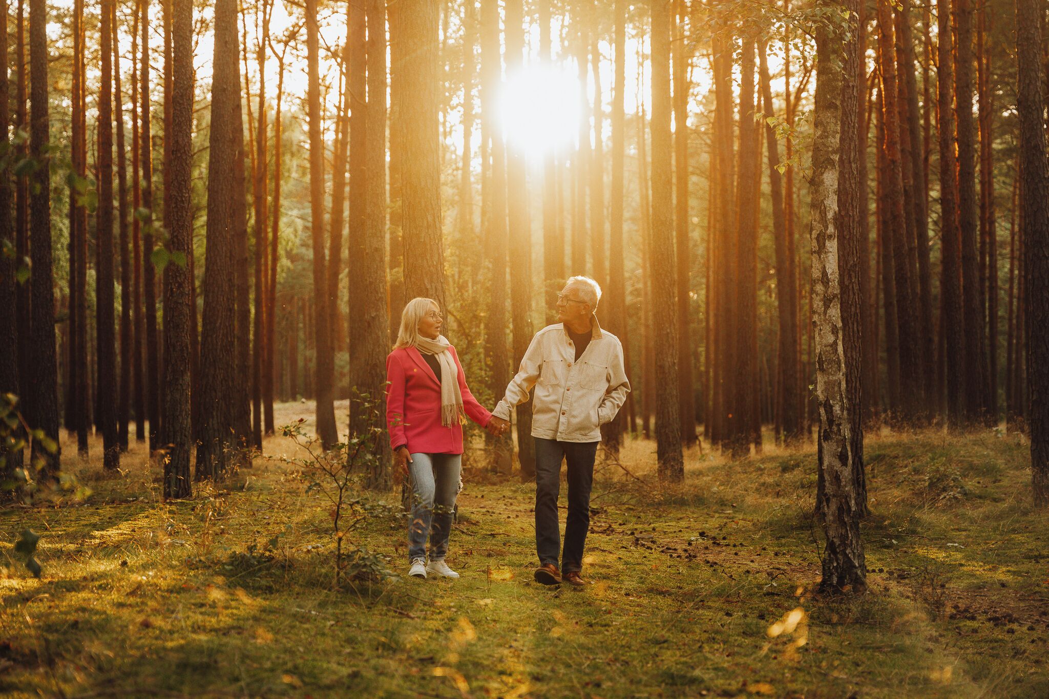 A couple holding hands in a forest with tall trees and sunlight filtering through.