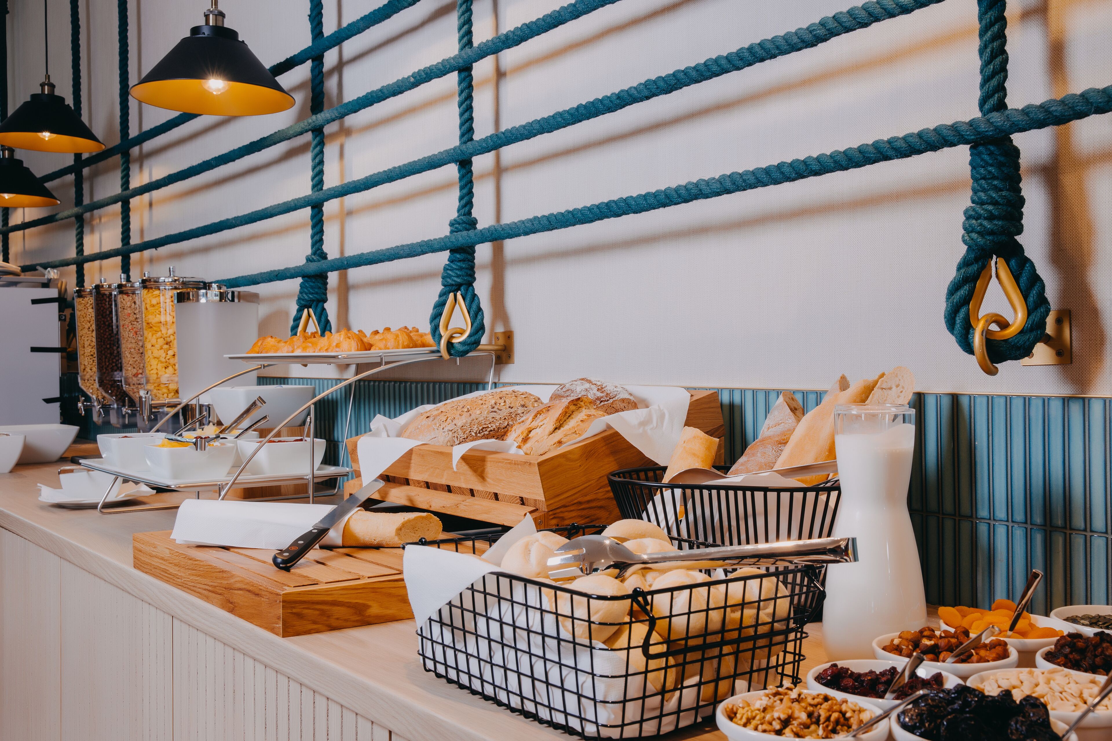 Ein reichhaltiges Buffet mit verschiedenen Speisen, darunter Brot, Gebäck und Beilagen, präsentiert auf einer Theke mit hängenden Seilen und Pendelleuchten.