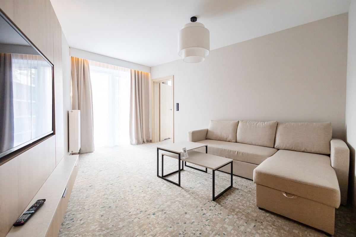 A modern hotel room with a sofa, a coffee table, a TV, and a large window with curtains.