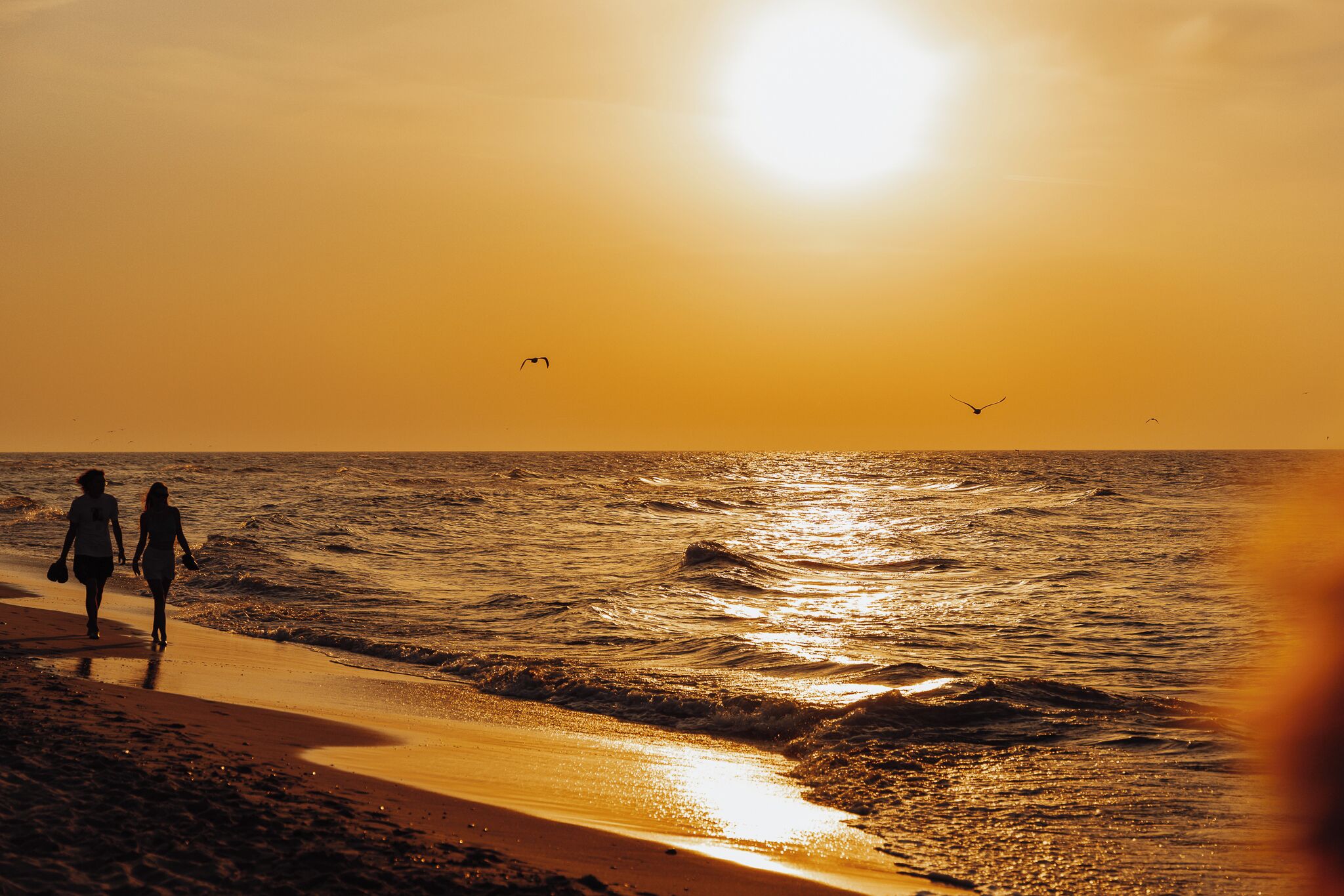 A beach scene with two people walking along the shore, the sun setting over the ocean.