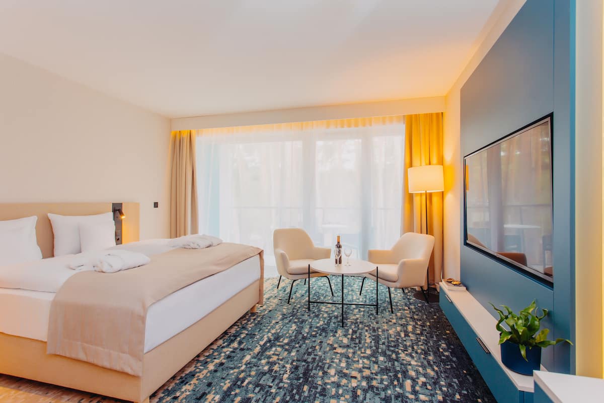 A hotel room with a bed, two chairs, a small table, a TV, and a plant.