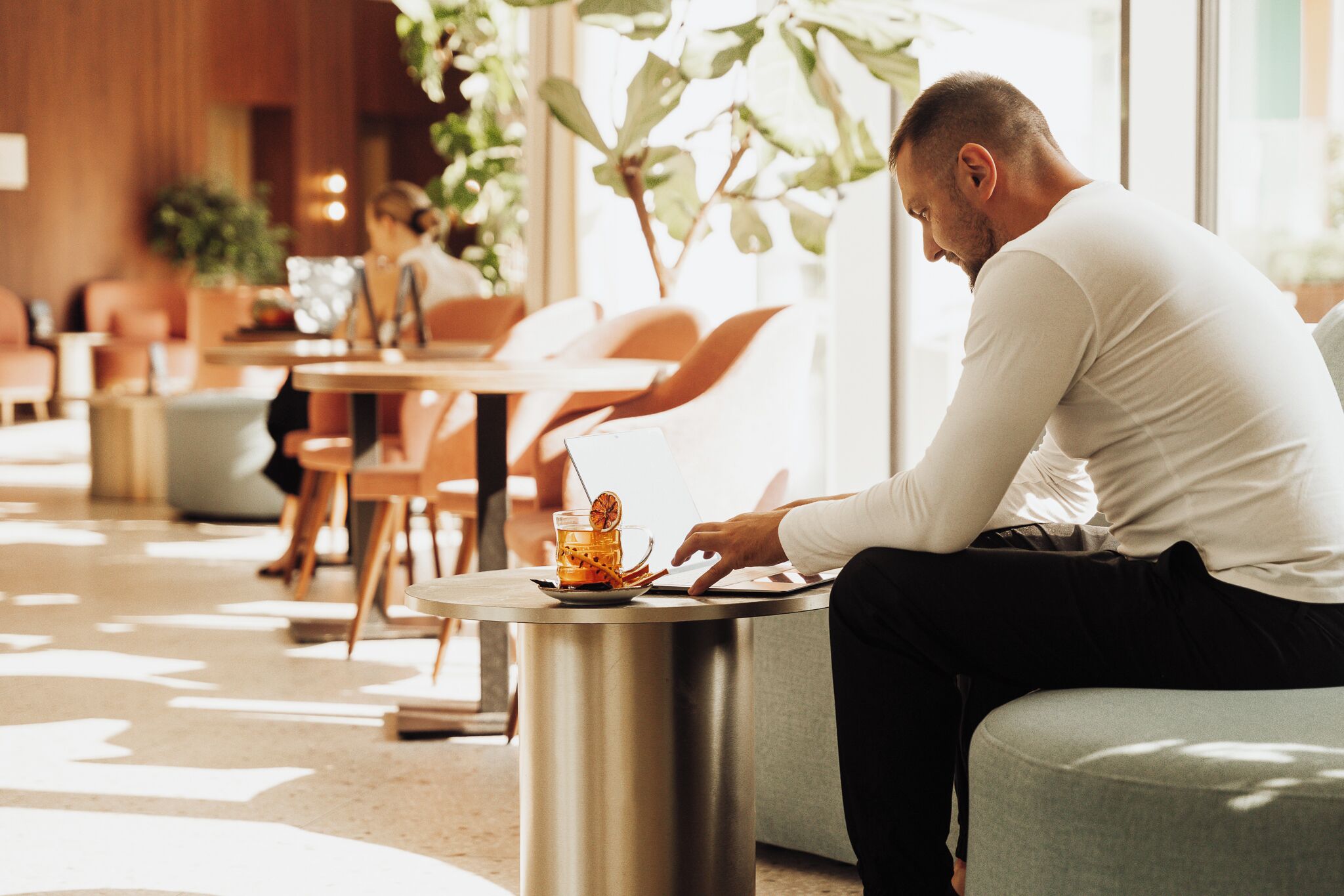 Un hombre sentado a una mesa con una bebida y una computadora portátil, en un espacio interior bien iluminado con muebles modernos y plantas.