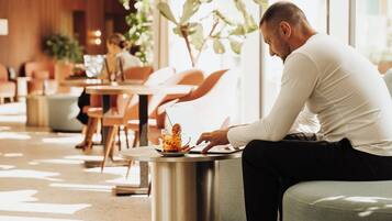 Un hombre sentado a una mesa con una bebida y una computadora portátil, en un espacio interior bien iluminado con muebles modernos y plantas.