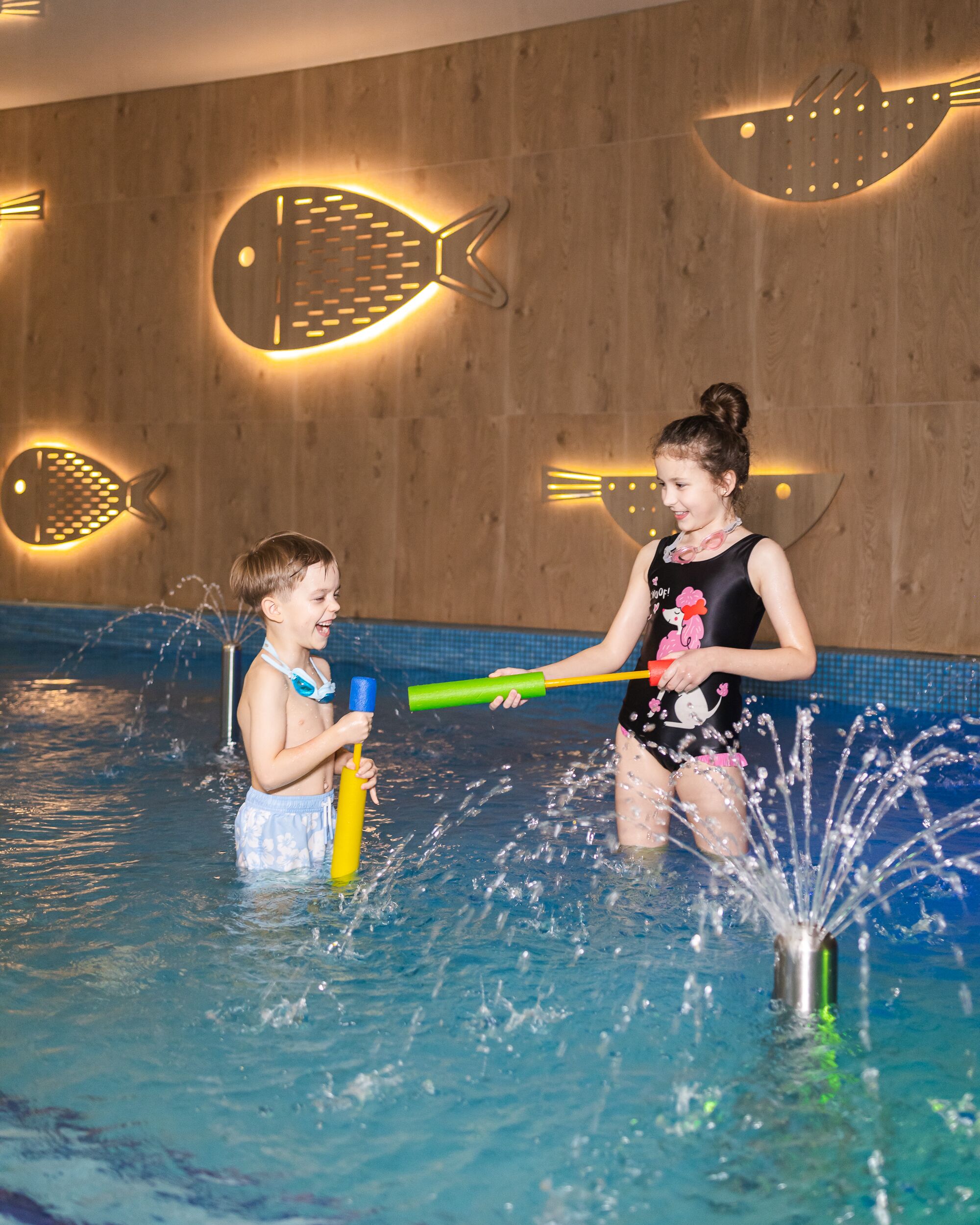 Two people in a swimming pool with water jets, wooden wall with fish-shaped lights, and a tiled floor.