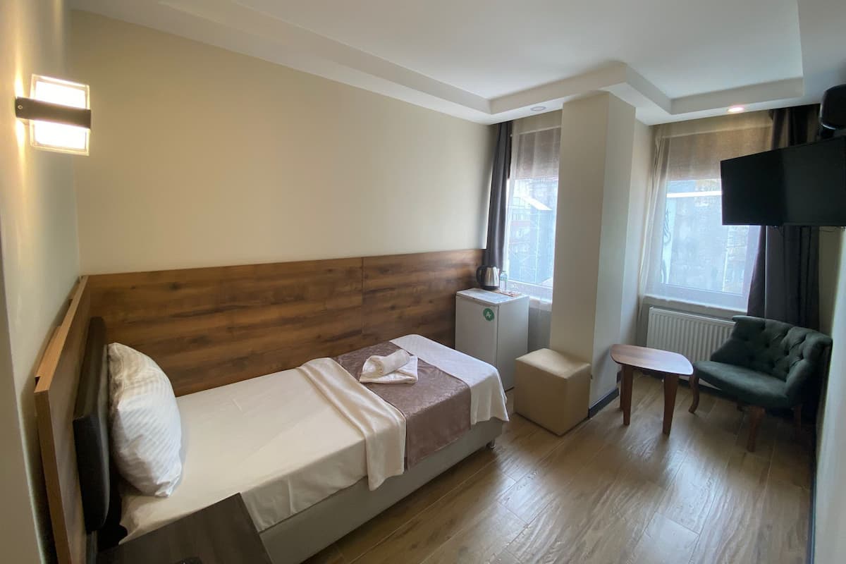 Habitación de hotel con cama de cabecera de madera, una mesita de noche pequeña, un sofá, un televisor y una ventana con cortinas.