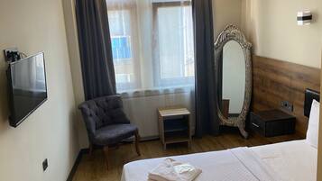 A hotel room with a bed, a TV, a chair, a window with curtains, a mirror, and a heater.