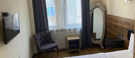 Una habitación de hotel con cama, televisión, silla, ventana con cortinas, espejo y calefactor.